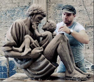 Timothy Scmaulz, while wearing a jeff cap, a watch, and a sleeveless shirt, working on a bronze sculpture which features Jesus as a young child who is being held in the lap of his father, Joseph, who has a beard. 