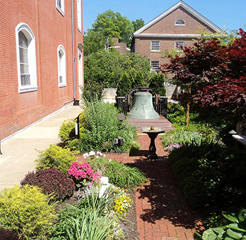 The Healing Garden next to Saint Augustine Parish - containing a large bell, a sundial, and flora while it is daytime. 